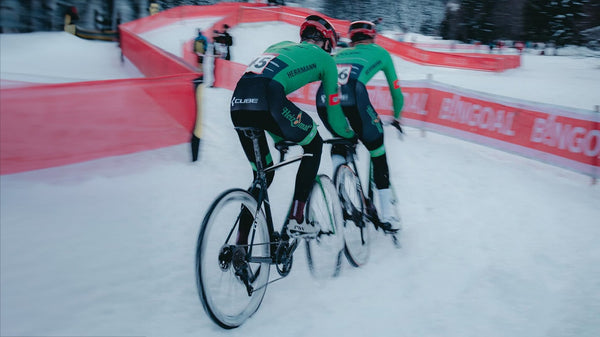 Two riders from the Heizomat powered by Kloster Kitchen Radteam ride through a race course covered in snow. 