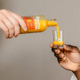 Contenu du kit de dégustation de shot de gingembre L : une personne verse un shot de gingembre curcuma dans un verre à shot à une autre personne.