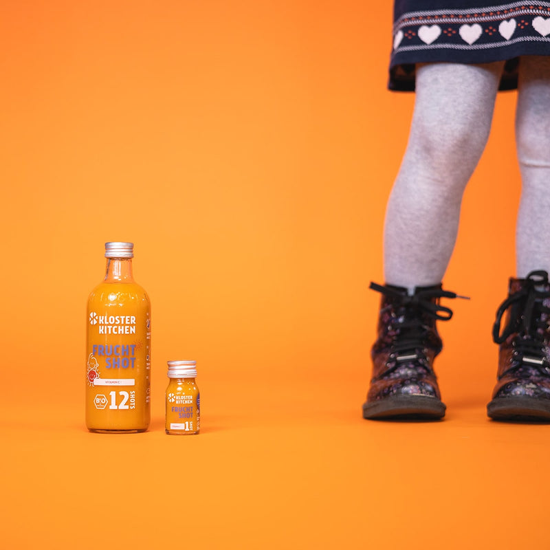 Contents of the fruit shot box: 1x fruit shot 12SHOTS in a 360 ml bottle, next to it 1x fruit shot 1SHOT in a 30 ml bottle. Next to it you can see children's legs wearing boots with flowers.