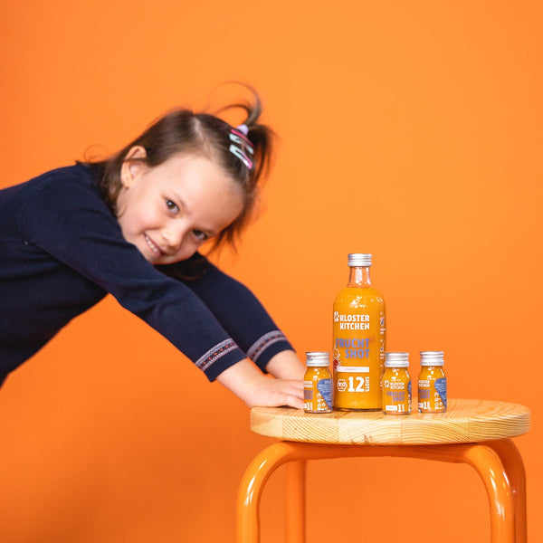 Contents of the fruit shot box: 1x fruit shot 12SHOTS bottle and 3x fruit shot 1SHOT on a stool. A little girl is leaning on the stool with both hands.