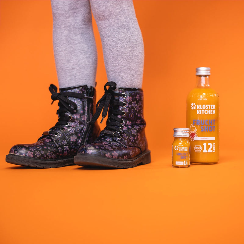 Contents of the fruit shot box: 1x fruit shot 12SHOTS in a 360 ml bottle, next to it 1x fruit shot 1SHOT in a 30 ml bottle. Next to it you can see children's legs wearing boots with flowers. 
