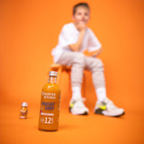 1x shot de fruits 12SHOTS bouteille de 360 ml. En arrière-plan, on voit flou 1x Fruit Shot 1SHOT dans une bouteille de 30 ml ainsi qu'un garçon assis à côté.