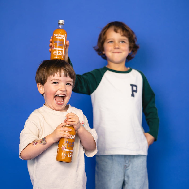 12SHOTS Frucht Shot dans une bouteille en verre de 360 ml est posé par un garçon sur la tête d'un autre garçon. Le garçon plus petit au premier plan, qui a la bouteille sur la tête, tient lui-même encore une bouteille de 12SHOTS Frucht Shot dans les mains.
