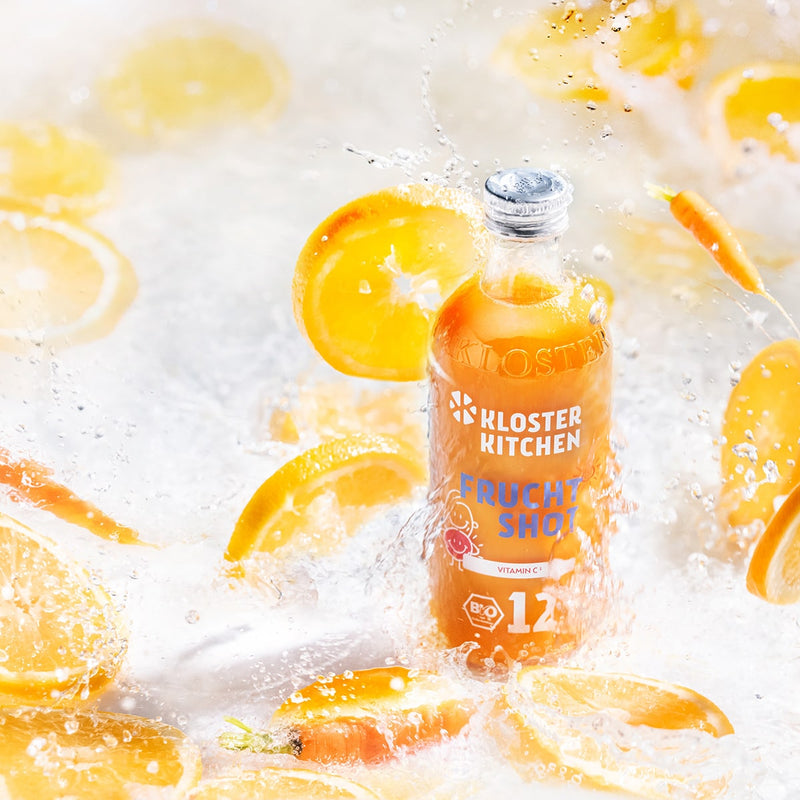 Fruit Shot 12SHOTS dans la bouteille de 360 ml se tient debout pendant que les tranches d'orange ainsi que la seringue d'eau passent devant elle.