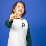 A boy has a fruit shot in his hand and holds it up to the camera while holding the lid of the bottle on his nose.