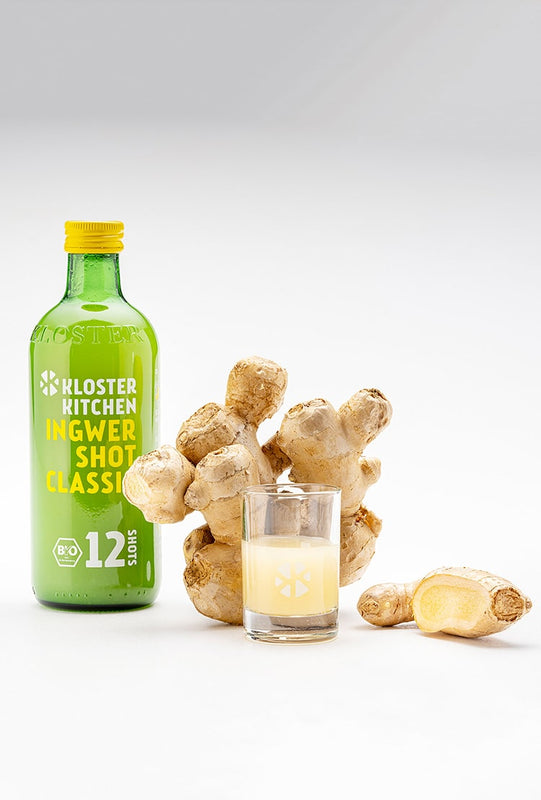 Un Ginger Shot Classic di Kloster Kitchen, accanto un bulbo di zenzero e uno shot versato in un bicchiere da shot.