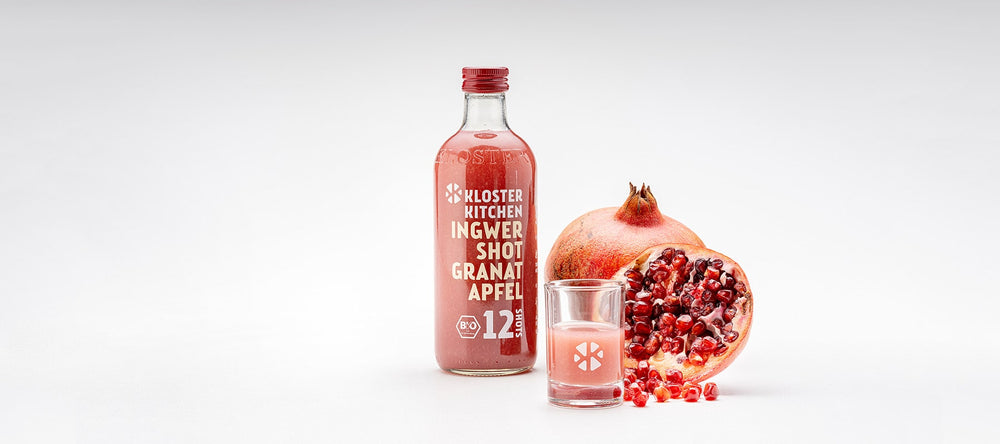 A ginger shot pomegranate 360 ml next to it a filled shot glass and behind it a sliced pomegranate and a still complete.