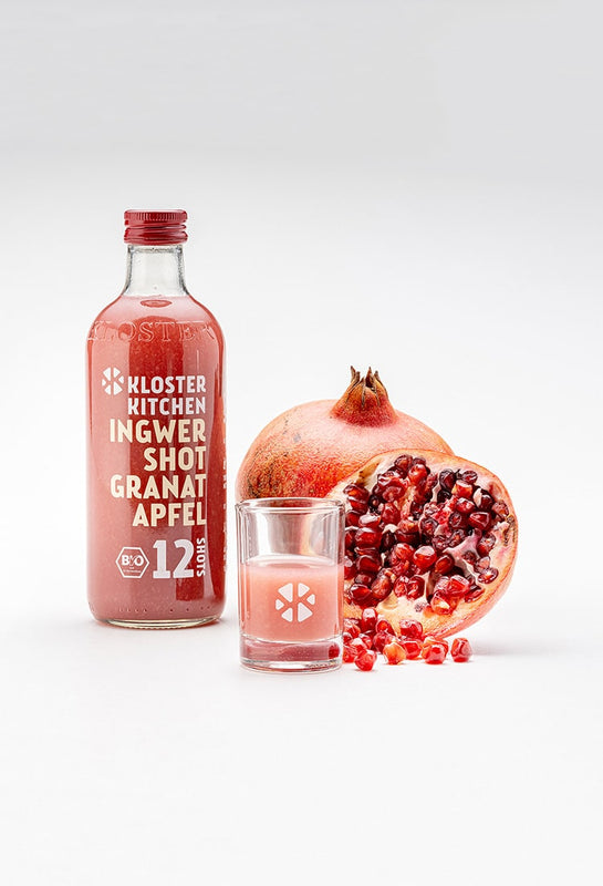 A ginger shot pomegranate 360 ml next to it a filled shot glass and behind it a sliced pomegranate and a still complete.
