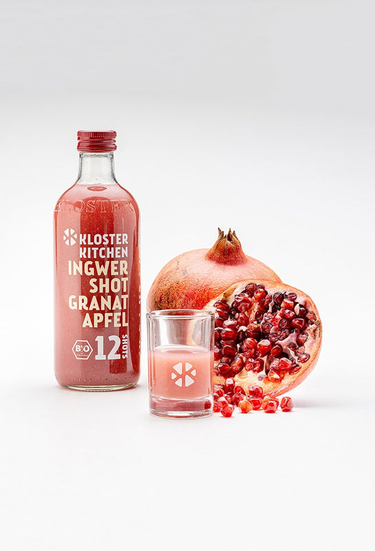 Ein Ingwer Shot Granatapfel 360 ml daneben ein gefülltes Shot Glas und dahinter ein aufgeschnittener Granatapfel sowie ein noch kompletter.