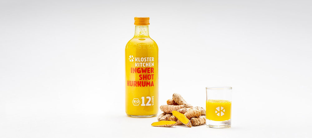 Le shot de gingembre au curcuma dans une bouteille de 360 ml, à côté de quelques morceaux de curcuma et à côté d'un verre à shot rempli de shot de gingembre au curcuma.