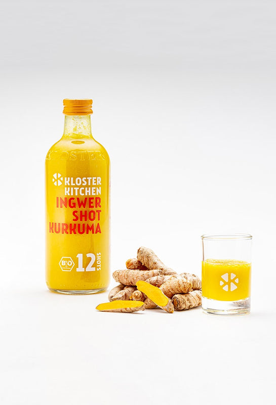 Le shot de gingembre au curcuma dans une bouteille de 360 ml, à côté de quelques morceaux de curcuma et à côté d'un verre à shot rempli de shot de gingembre au curcuma.