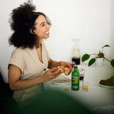 1 of the 3x 12SHOTS 360 ml Ginger Shot Classic: A Ginger Shot Classic 12SHOTS stands on a table where a woman is sitting and just eating a croissant. Next to the bottle is a filled shot glass full of Ginger Shot, ready to be drunk.