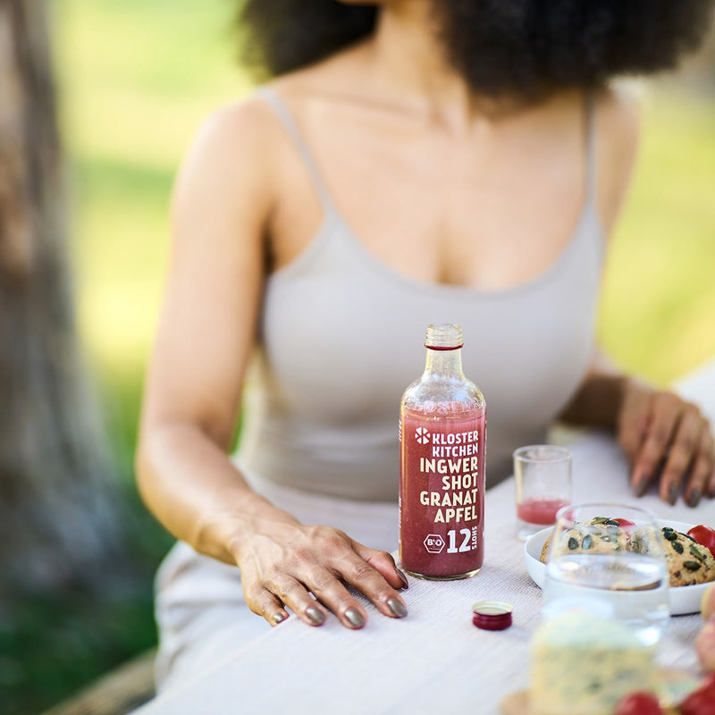 Contents Ginger Shot Tasting Set M: Ginger Shot Pomegranate 360 ml bottle stands on a laid table, you can see a woman who poured the shot into a shot glass. 