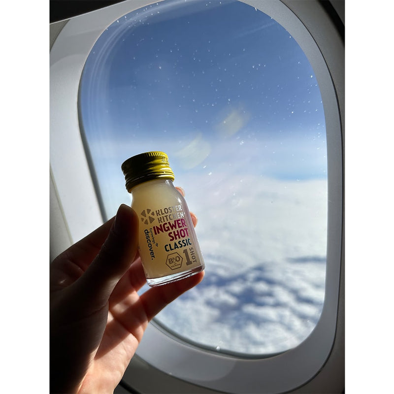 The Ginger Shot Classic is held up to a window of an airplane. Outside the window you can see a cloud cover.