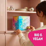 Kloster Kitchen Adventskalender in bunter Faltschachtel mit Pink-, Blau- und Gelbtönen, Bio-Siegel und nummerierten Türchen; eine Person öffnet ein Türchen und hält ein kleines gelbes Produkt in der Hand.