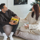 (Paar mit Geschenkbox): Mann und Frau sitzen gemütlich vor dem Weihnachtsbaum und übergeben sich eine Kloster Kitchen Geschenkbox mit gelber Schleife