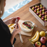 Miras por encima del hombro de una mujer que sostiene un Ginger Shot Granada en un frasco de 30 ml. Sobre la mesa puedes ver un cuenco que ella ha preparado, un bol lleno de fruta y el envase abierto de la cura Ginger Shot.