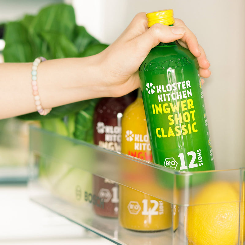 The contents of a Ginger Shot Mix Box from Kloster Kitchen in a refrigerator compartment. One hand is reaching for the Ginger Shot Classic bottle.