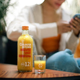 1 von den 3x 12SHOTS 360 ml Ingwer Shot Kurkuma steht auf einem Beistelltischchen. Daneben steht ein gefülltes Shot Glas. Im Hintergrund sieht man jemand auf einer Couch sitzen.