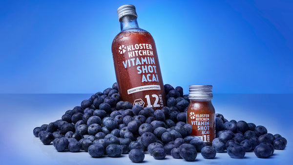 Il nuovo Vitamin Shot Acai di Kloster Kitchen - appena lanciato! Una bottiglia da 360 ml e una da 30 ml una accanto all'altra in un mucchio di bacche di acai.