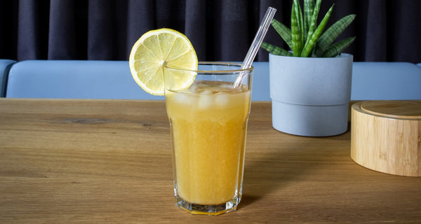 Ginger shot pineapple iced tea in a glass with a slice of lemon tucked into the rim as decoration. There is also a glass straw tucked into the glass.