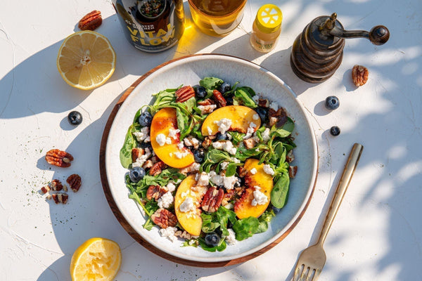 Une salade avec des pêches, des noix, des myrtilles et de la feta en topping, encadrée de citrons, d'un moulin à poivre, d'un shot de gingembre Classic et de Vital Flow