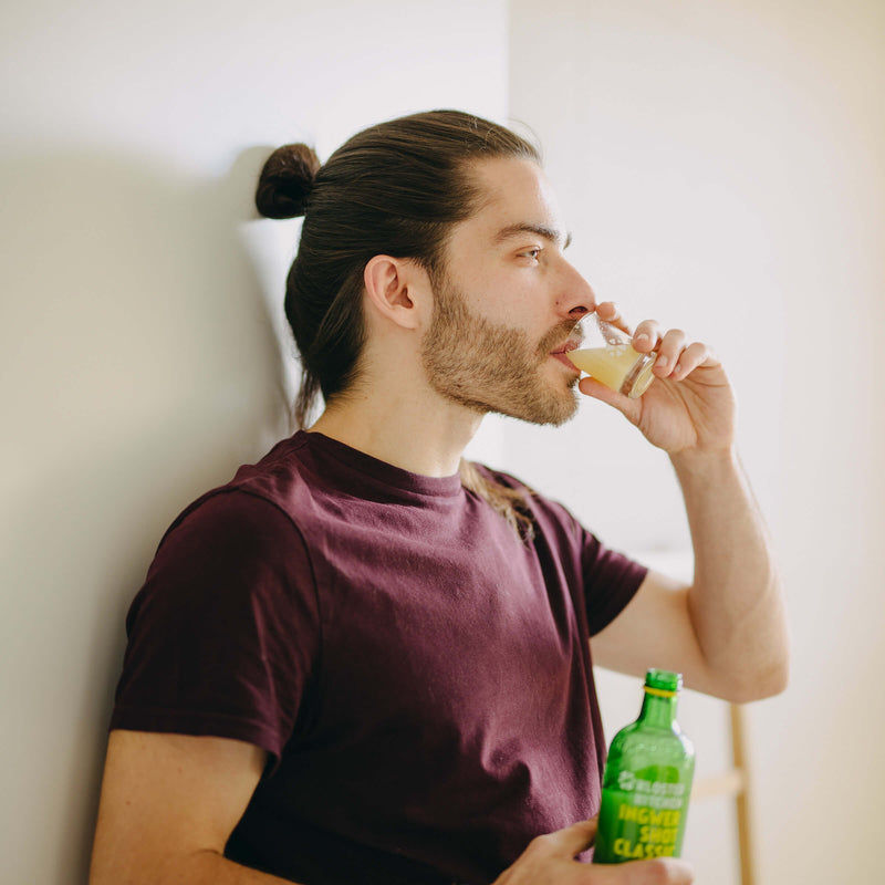 Gläser für Ingwer Shots: Mann lehnt an der Wand und hat eine Flasche Ingwer Shot Classic in der Hand, während er aus einem vollen Shot Glas für Ingwer Shots trinkt.