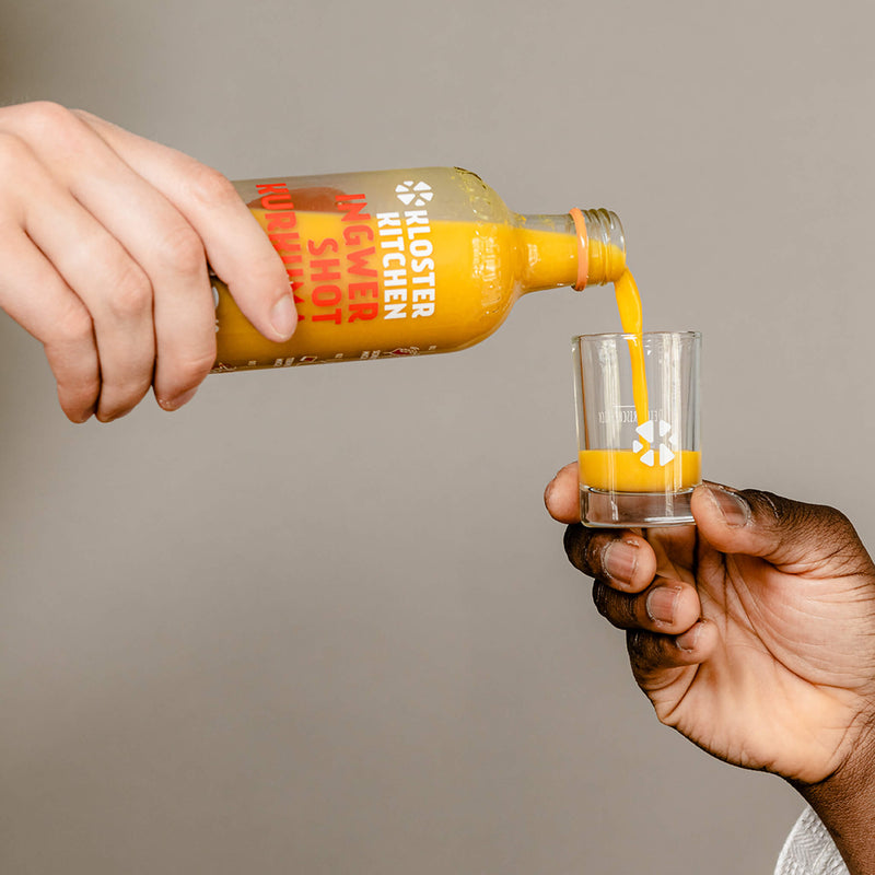 Contents of Ginger Shot Tasting Set L: One person pours a Ginger Shot Turmeric into a shot glass for another.