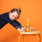 Contenu de la boîte de shots de fruits : 1x bouteille de shots de fruits 12SHOTS et 3x shots de fruits 1SHOT sur un tabouret. Une petite fille s'appuie des deux mains sur le tabouret.