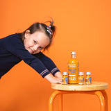 Contenuto della scatola di shot di frutta: 1x bottiglia di shot di frutta 12SHOTS e 3x shot di frutta 1SHOT su uno sgabello. Una bambina si appoggia allo sgabello con entrambe le mani.