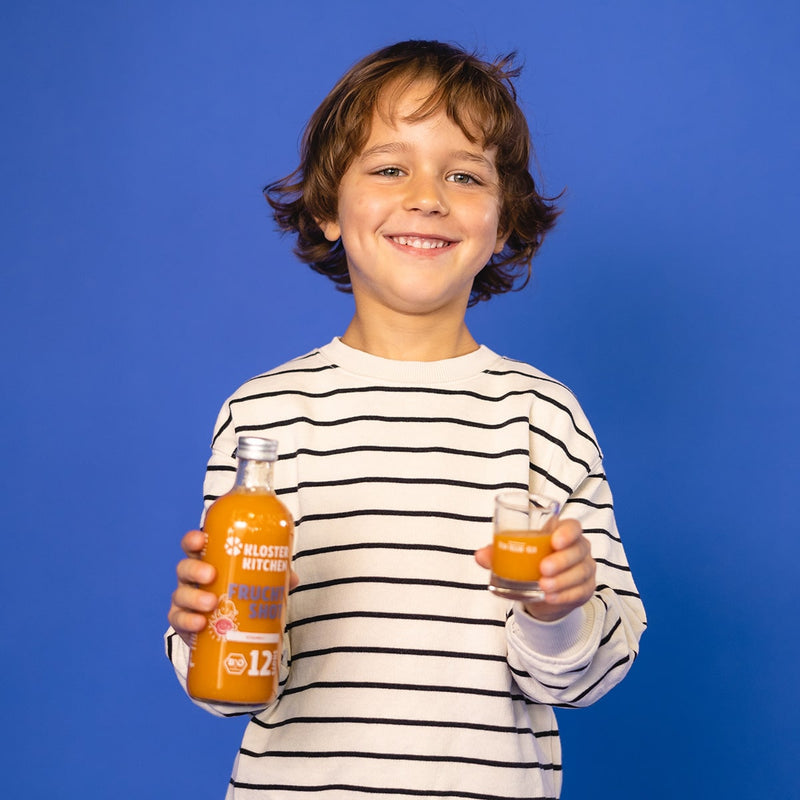 Un petit garçon tient dans sa main le Fruch Shot 12SHOTS dans une bouteille en verre de 360 ml ainsi qu'un shot versé dans un verre à shot.