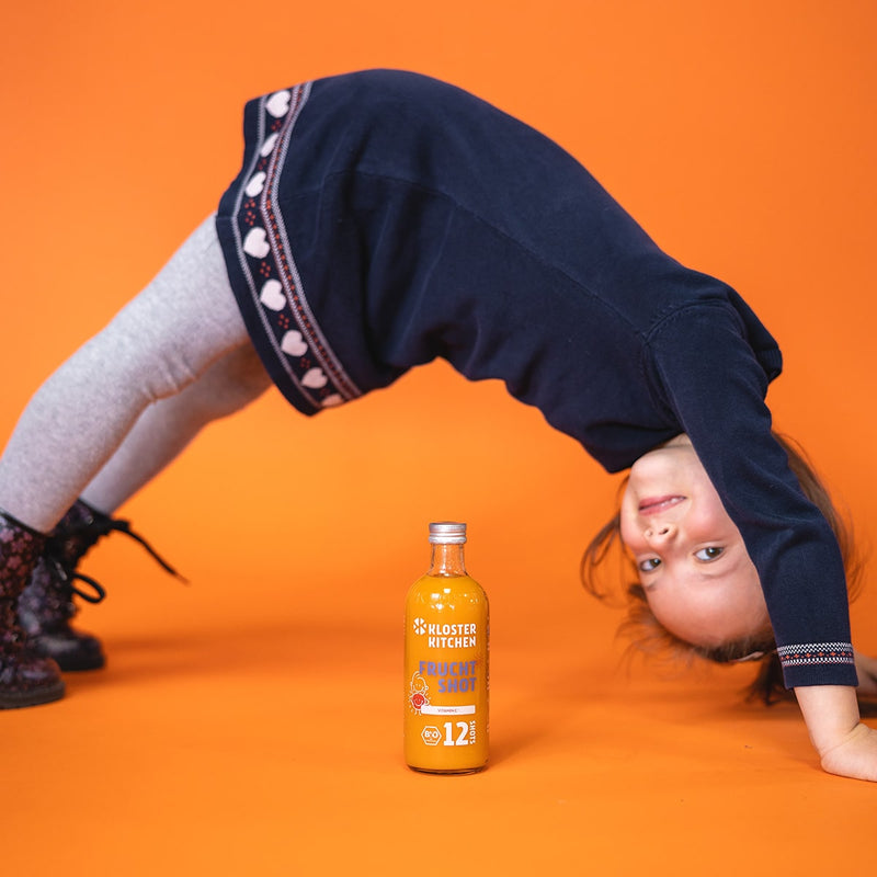 Une petite fille, debout, a les mains sur le sol et forme pour ainsi dire un toit au-dessus d'une bouteille de 12SHOTS Frucht Shot 360 ml.