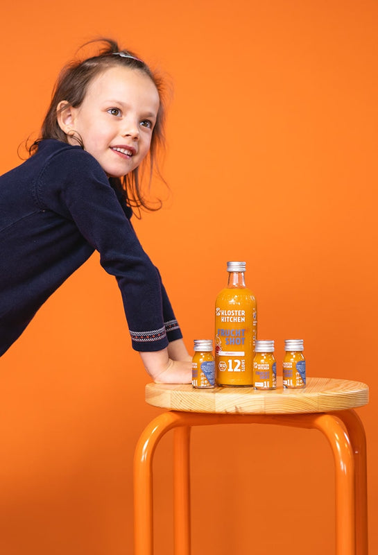 Mädchen stützt sich an einem Hocker ab, auf dem Frucht Shots stehen in der großen 360 ml Flasche sowie daneben drei Frucht Shots in 30 ml Fläschchen. 