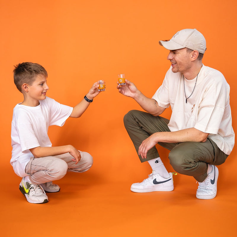 An adult and a child kneel opposite each other and toast each other with a fruit shot in a 30 ml bottle.
