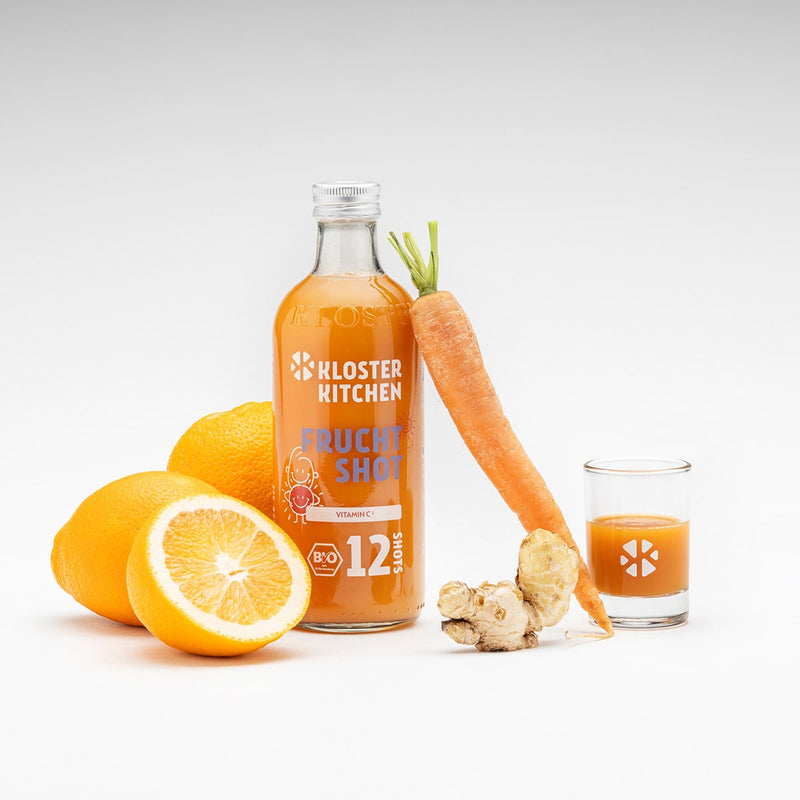 Un shot de fruits dans une bouteille de 360 ml et un verre à shot rempli avec un shot versé. Une carotte est appuyée contre la bouteille, ainsi qu'un tubercule de gingembre et quelques oranges.