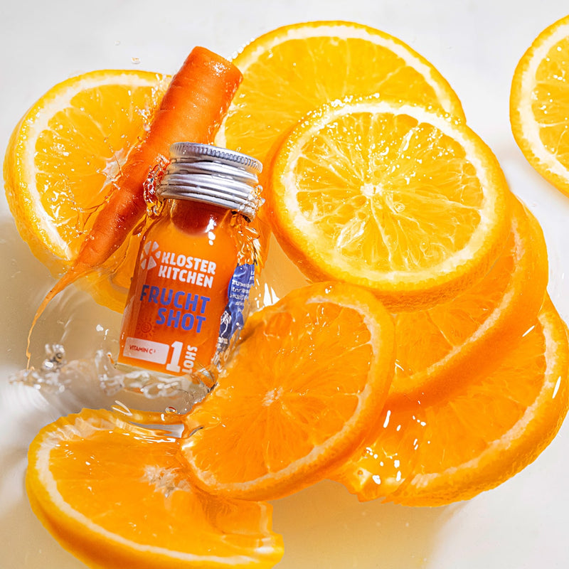 A fruit shot in a 30 ml bottle falls into crystal-clear water. There are also lots of orange slices and a small carrot floating in the water. 