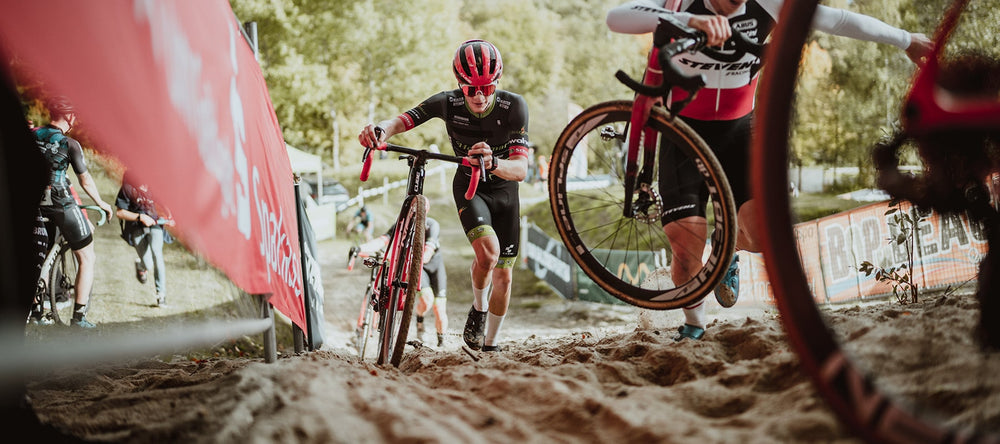 Team member of the Heizomat cycling team powered by Kloster Kitchen pushes his bike up a steep sandy hill. In front and behind him are other riders.