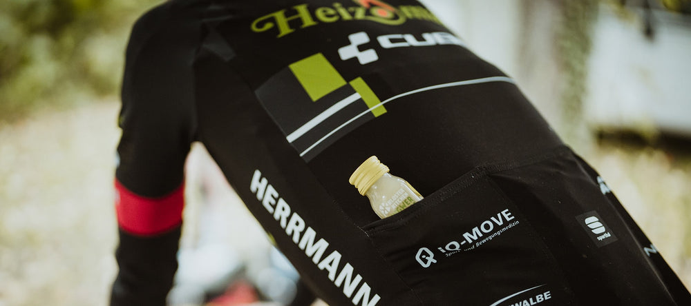 Nahaufnahme des Rückens eines Fahrers des Heizomat Radteams powered by Kloster Kitchen. Man erkennt den Fahrer an seinem Heizomat Trikot. In seiner Rückentasche steckt ein Ingwer Shot von Kloster Kitchen.