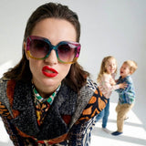 A woman wearing large, colorful sunglasses and red lipstick looks directly into the camera, while two small children play together and laugh in the background. The scene focuses on the stylish mother in the foreground and the lively family scene in the background—perfect for Mother’s Day.