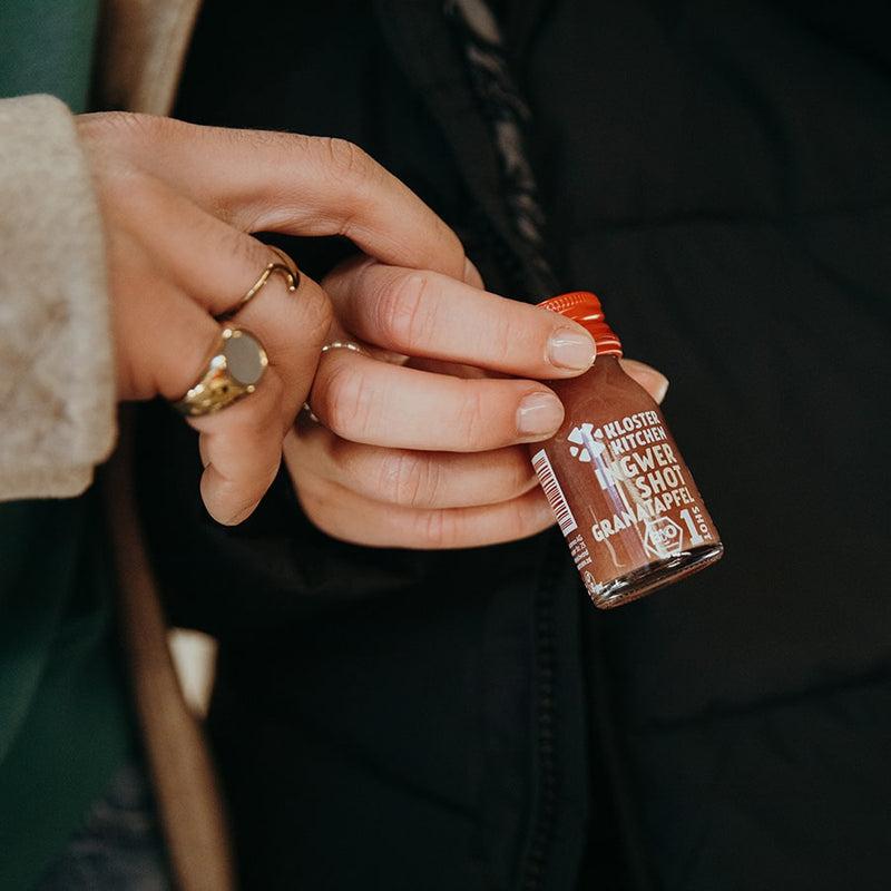 Contents of the Kloster Kitchen gift box Mother's Day: one hand holds a ginger shot pomegranate 30 ml bottle, the hand of a second person meanwhile holds on to the first hand with two fingers.  