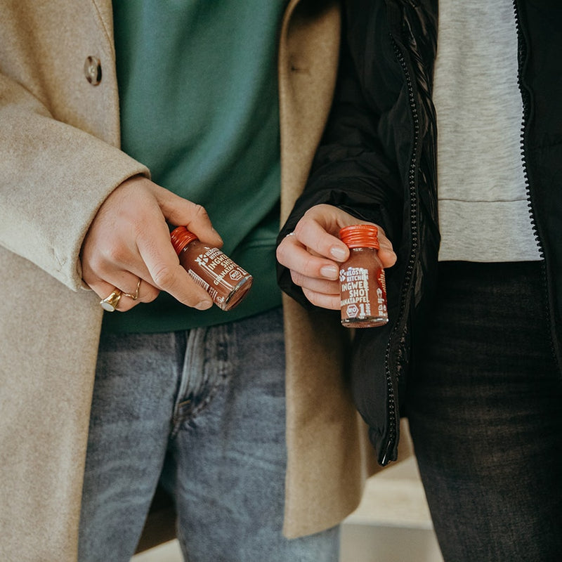 Contents of Kloster Kitchen gift box Valentine's Day. Two people each holding a ginger shot pomegranate 30 ml bottle.