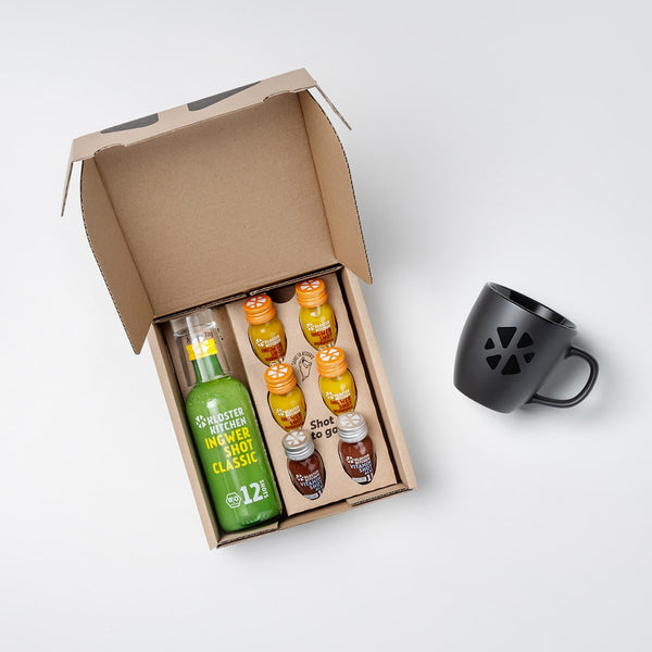 Hot Shot Box from Kloster Kitchen: An opened box with a 360 ml bottle of Ginger Shot Classic, next to it small 30 ml bottles with 4x Ginger Shot Turmeric and 2x Vitamin Shot Acai. Next to the opened box is a matt black cup with the Kloster Kitchen logo on it