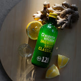 Inhalt des Probierset M: Ingwer Shot Classic Flasche liegeng auf einem Holzbrett, daneben ein Shot Glas, aufgeschnittene Zitronenstücke und eine Ingwerknolle