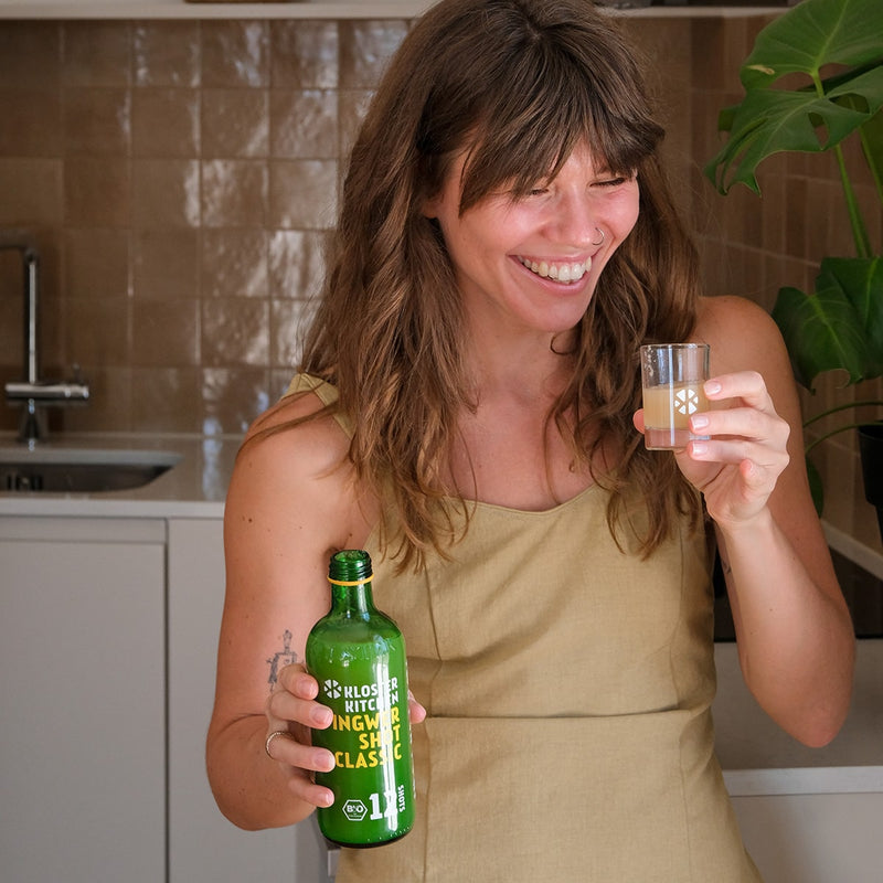 Inhalt des Probierset S: Frau trinkt einen Ingwer Shot Classic aus einem Shot Glas und hält die Flasche mit der anderen Hand.