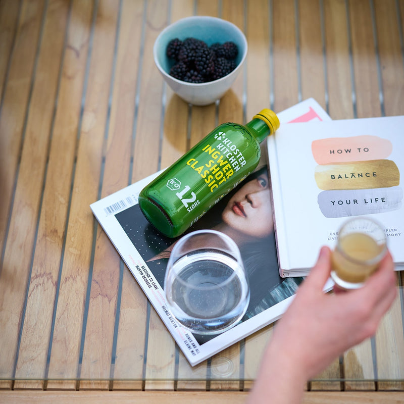 Der Inhalt einer Ingwer Shot Mix Box von Kloster Kitchen: Der Ingwer Shot Classic 12SHOTS liegt auf einem Tisch zwischen Zeitschriften, einem Glas und einer Schale voller Brombeeren.