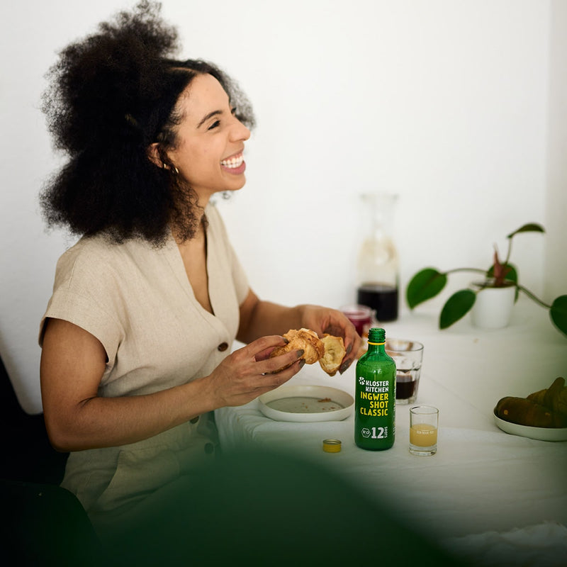 1 von den 3x 12SHOTS 360 ml Ingwer Shot Classic: Ein Ingwer Shot Classic 12SHOTS steht auf einem Tisch, an dem eine Frau sitzt und gerade ein Croissant isst. Neben der Flasche  steht ein gefülltes Shot Glas voller Ingwer Shot, fertig, um getrunken zu werden.