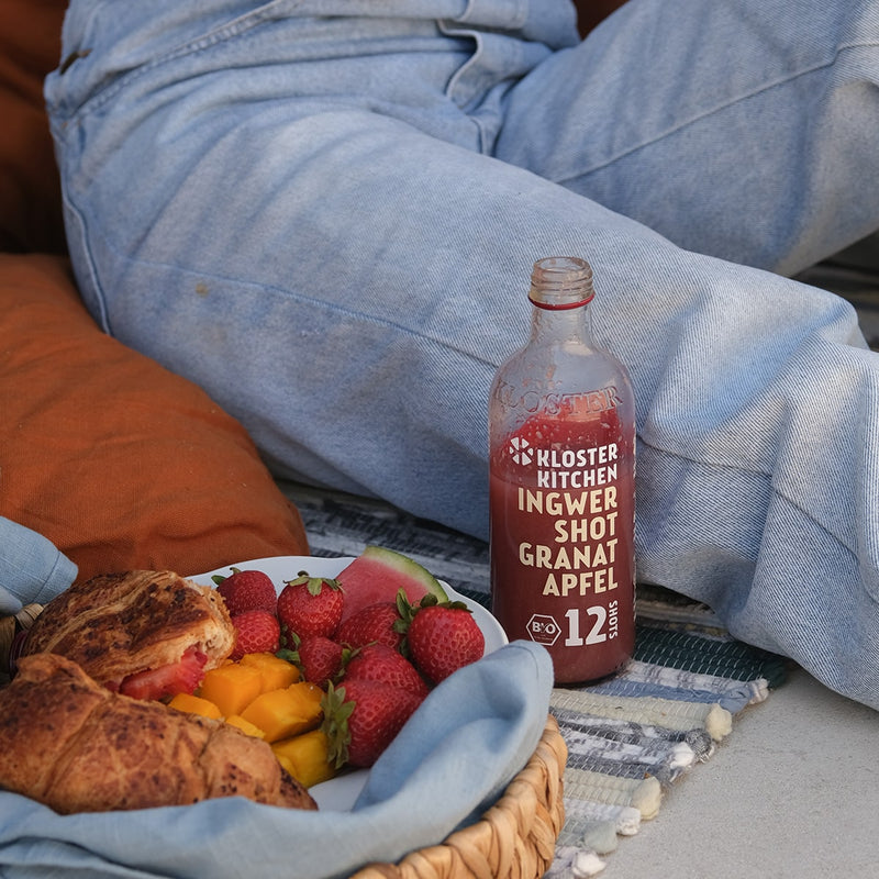 1 von den 3x 12SHOTS 360 ml Ingwer Shot Granatapfel: Der Ingwer Shot Granatapfel 12SHOTS steht auf einer Picknick Decke, neben einem Korb mit Früchten und Gebäck.