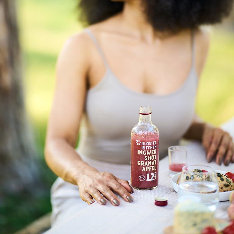 Contenuto del set di degustazione Ginger Shot M: la bottiglia Ginger Shot Pomegranate da 360 ml si trova su un tavolo apparecchiato, si vede una donna che versa lo shot in un bicchierino. 