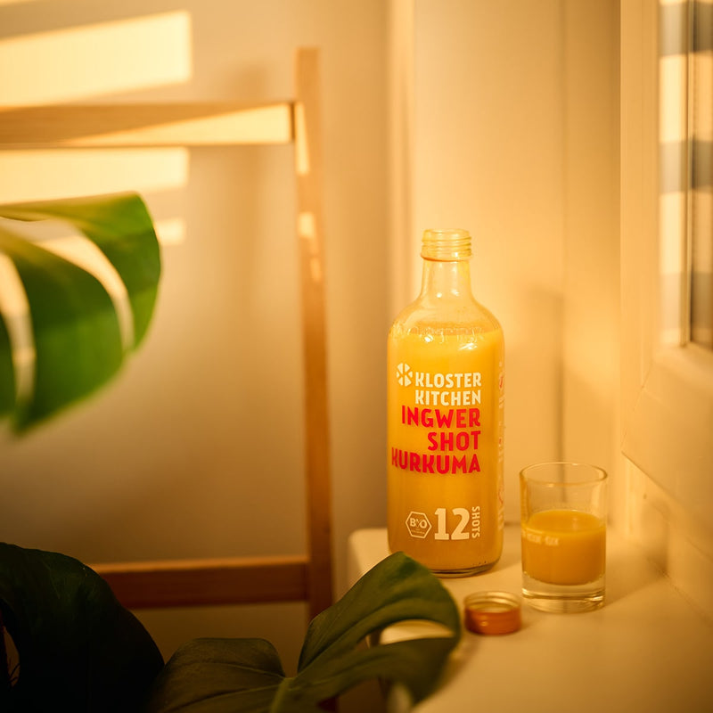 1 von den 3x 12SHOTS 360 ml Ingwer Shot Kurkuma steht auf einem Fenstersims, daneben steht ein gefülltes Shot Glas.