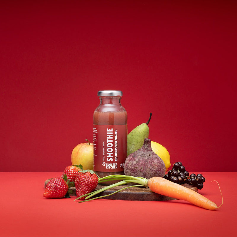 Smoothie mela fragola pera su sfondo rosso. Il frullato si trova su una tavola di legno e gli ingredienti sono allineati accanto alla bottiglia: fragole, mela, barbabietola, pera, limone, ribes rosso e una carota.